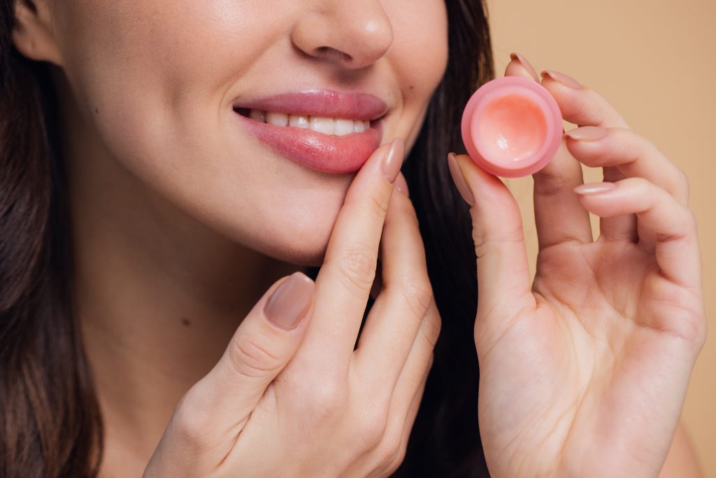 Lip Care Routine. Close up beauty portrait of smiling woman cares of her lips on beige background. Young brunette woman applying make-up cosmetic SPA product. Model using lip balm or gloss for hydration, nutrition, smoothing balm.