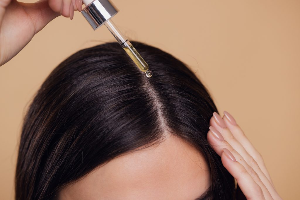 Scalp Care Routine. Close up view of unrecognizable young woman putting hair serum for alopecia and hair growth in beige background with selective focus