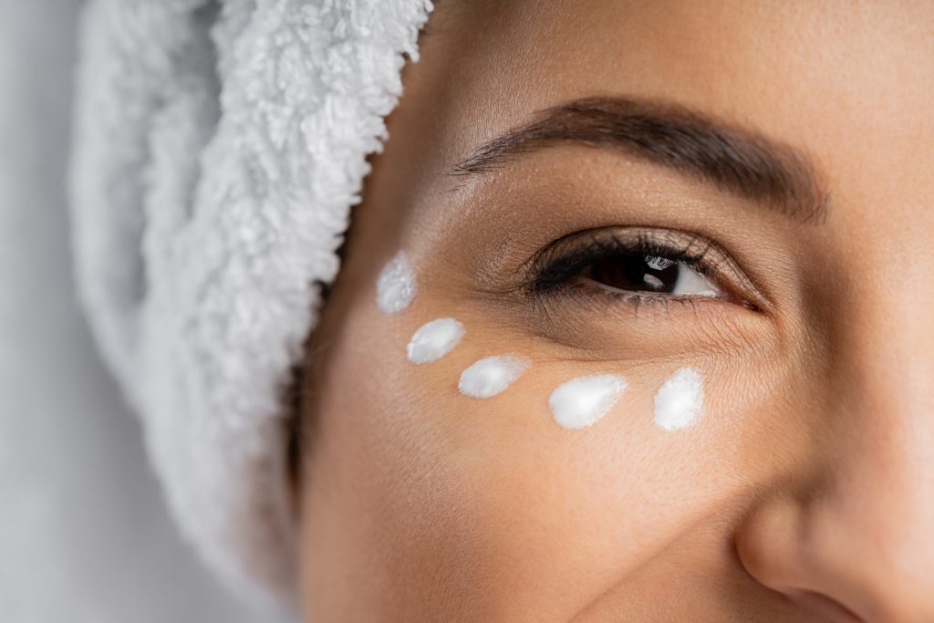 Under Eye Cream. Cropped view of woman with towel and cream under eye isolated on grey