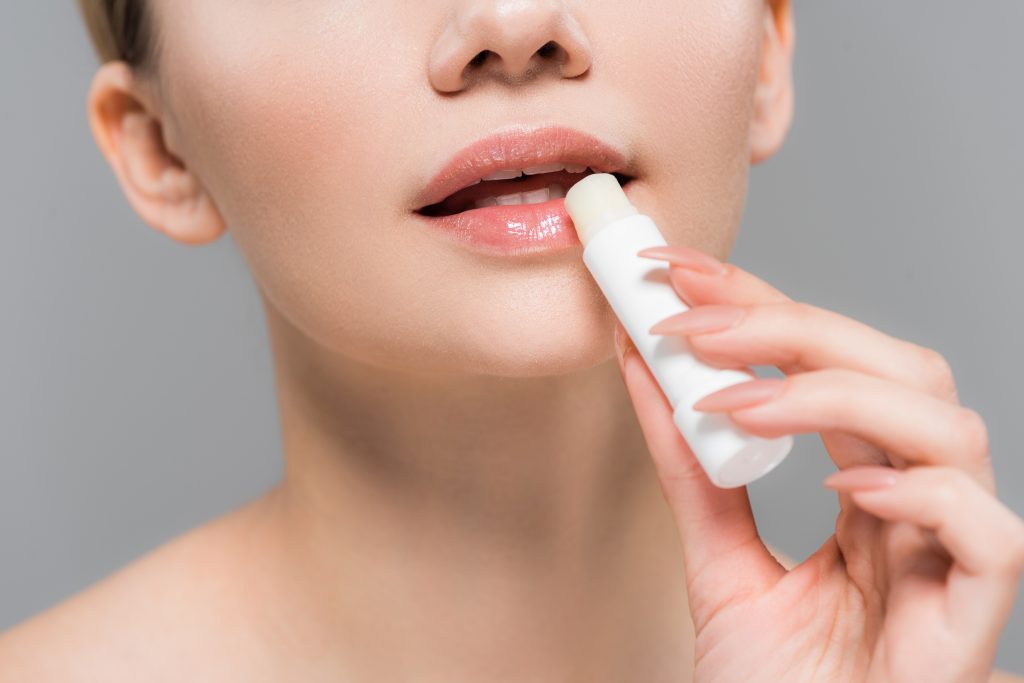 cropped view of young woman holding lip balm near lips isolated on grey