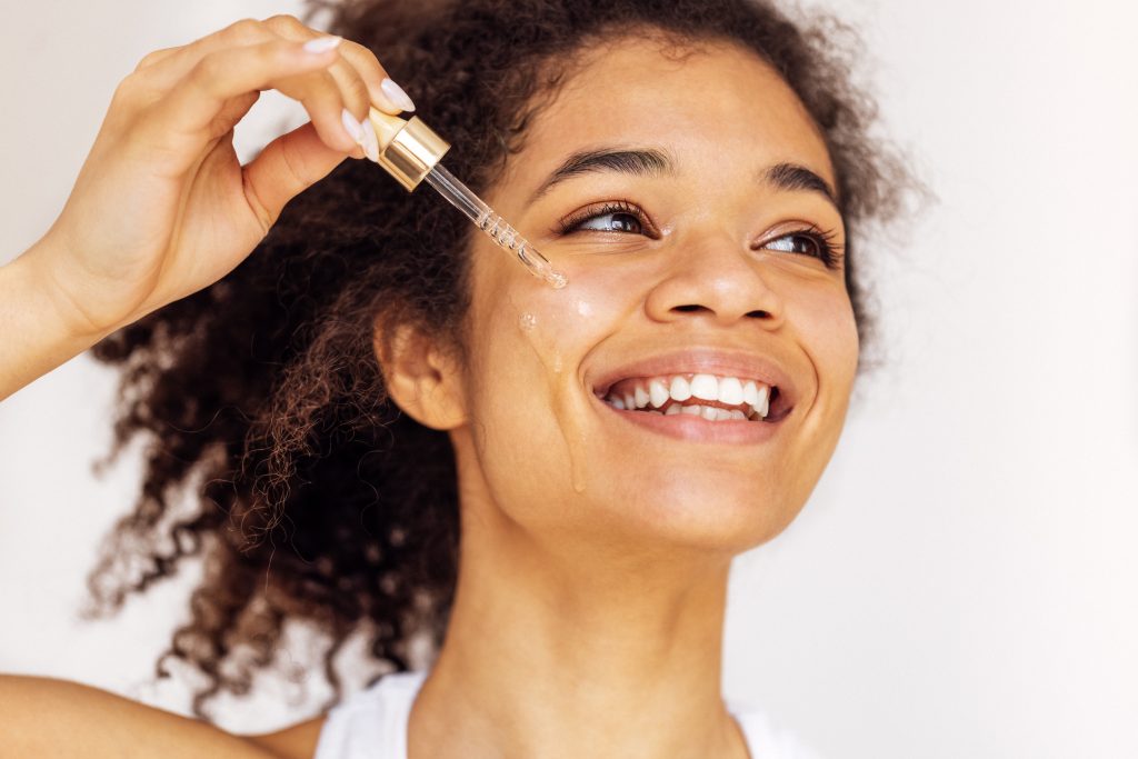 Cute smiling african young woman applies moisturizing serum from pipette to her cheek. Lovely charming mixed race girl taking care of her facial skin. Cosmetics and beauty concept. face serum. facial care. skincare.