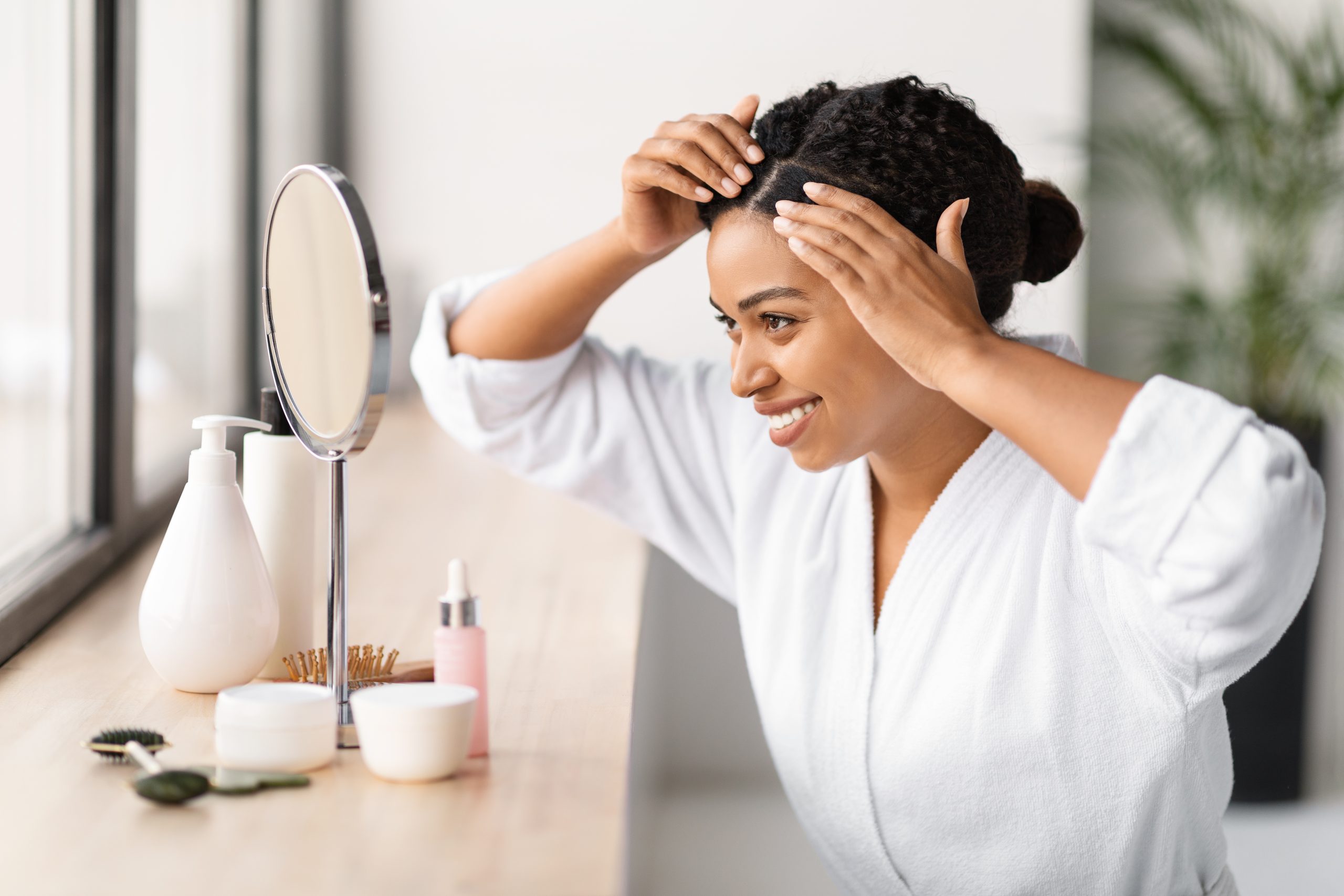 No Dandruff. Cheerful young black woman in bathrobe styling her hair while looking in vanity mirror in bathroom, beautiful african american female checking hair roots and smiling, closeup.