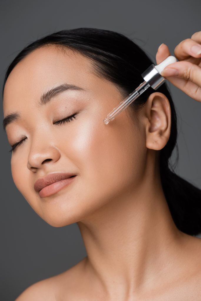 young asian woman with closed eyes applying cosmetic serum on clean face isolated on grey. niacinamide for skin.