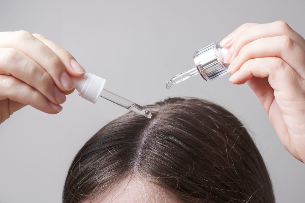 woman with beautiful hair, applies vitamins for hair growth. Young woman applying oil to her hair with a pipette on a gray background close up.