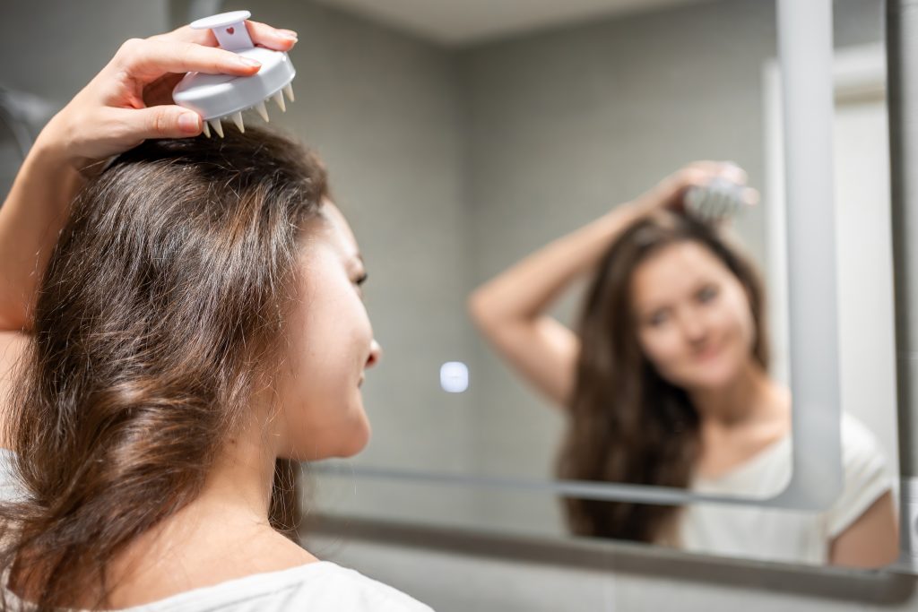 Young woman doing self hair scalp massage with scalp massager or hair brush for hair growth stimulating at home. Reflected view of the mirror. High quality photo