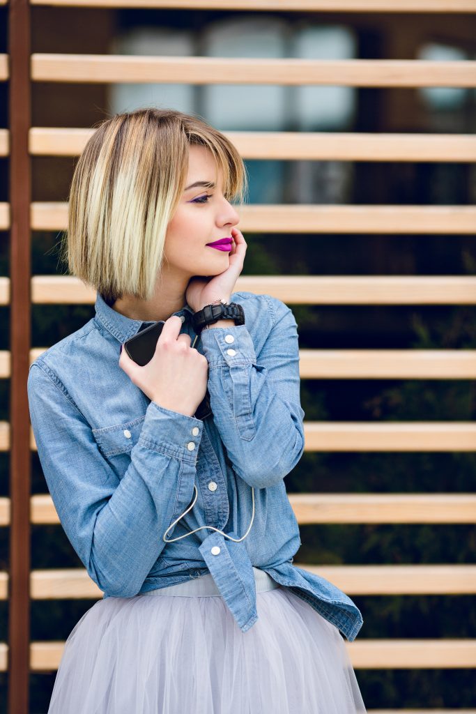 A close portrait of a dreamy girl with bright pink lips and short blond hair looking to her left listening to music on a smartphone with striped wooden balks behind wearing blue jeans shirt and grey tulle skirt. Timeless Haircuts for Fine Hair.