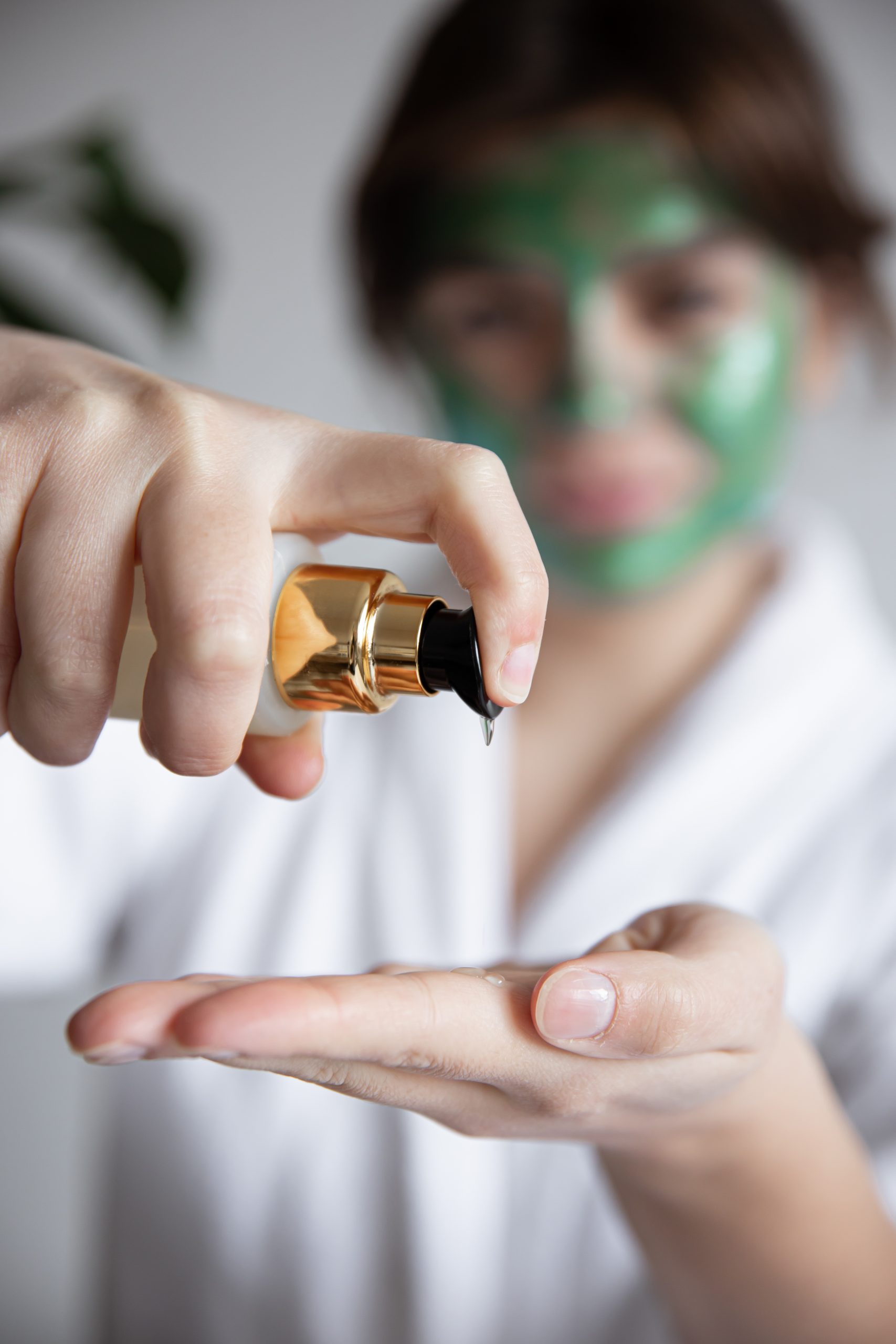 Slugging Skincare. Close-up, a woman in a white robe with a green cosmetic mask presses on the dispenser of beauty care products, blurred background. Skincare. face care.