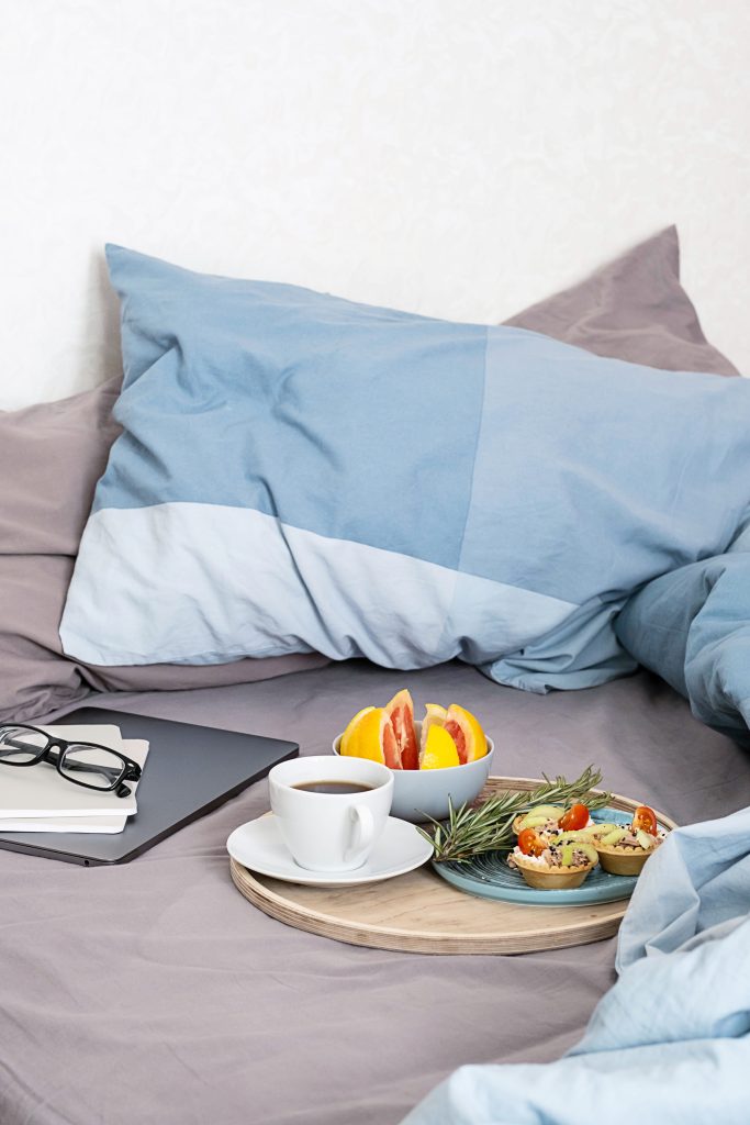 Coffee cup, cold tartlets and grapefruit on wooden tray, laptop, eyeglass and notebook on bedding. Breakfast in bed, cosy morning at home.