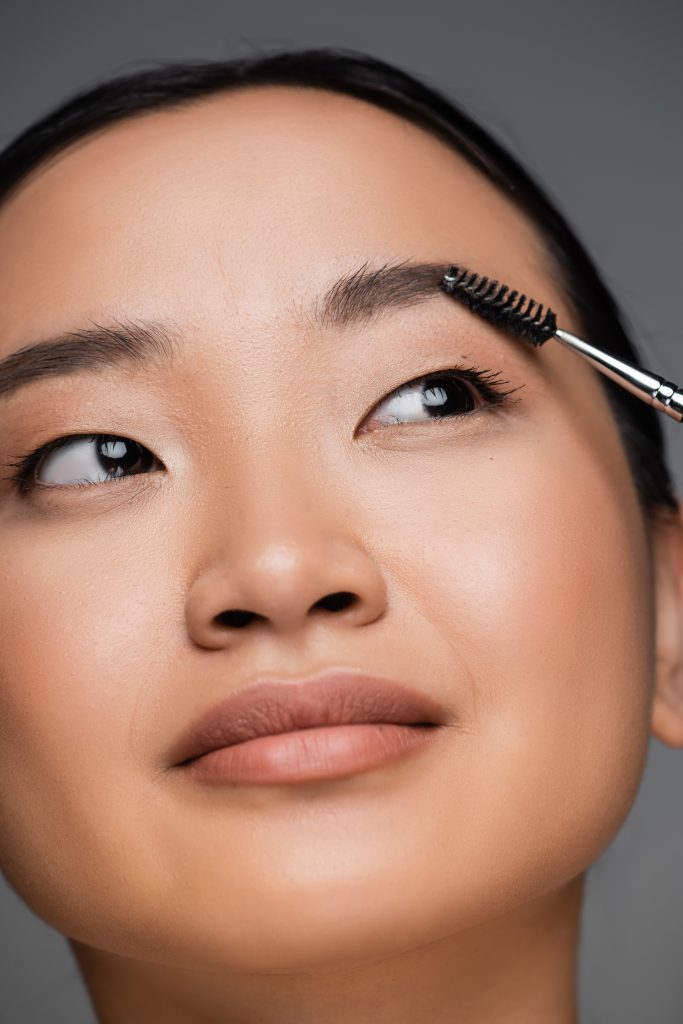 Eyebrow Growth Serums. close up portrait of pretty asian woman brushing eyebrow isolated on grey. 