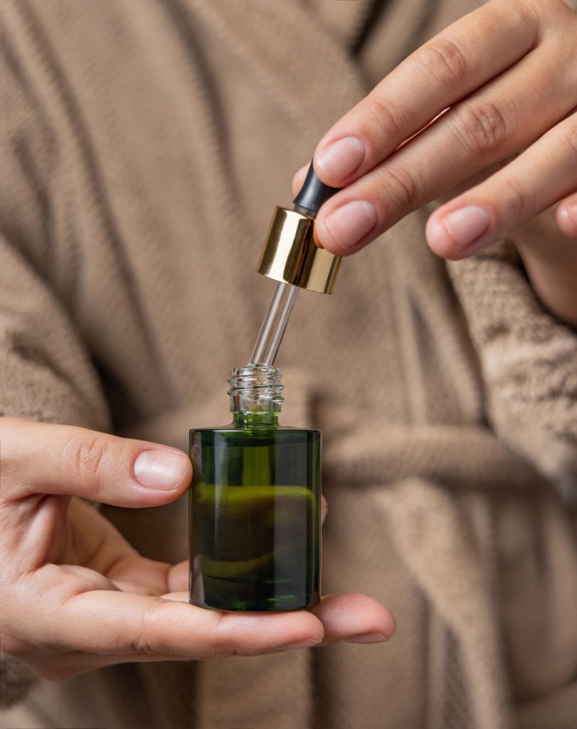 Female hands with natural manicure opening white green cosmetic dropper bottle. Close up of woman in bathroom wearing light brown bathrobe with drop of serum or essence falling from pipette