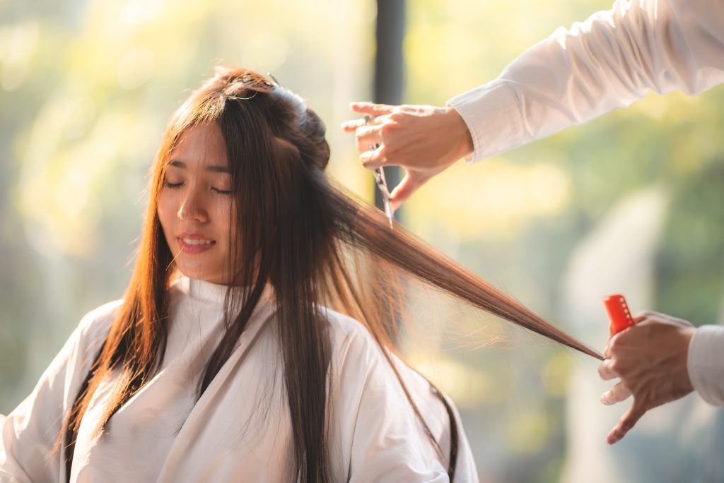 Young beautiful and smiling woman sitting in salon chair with cloth wrapped around getting hair cut and hair styling done in modern salon by experienced male professional barber