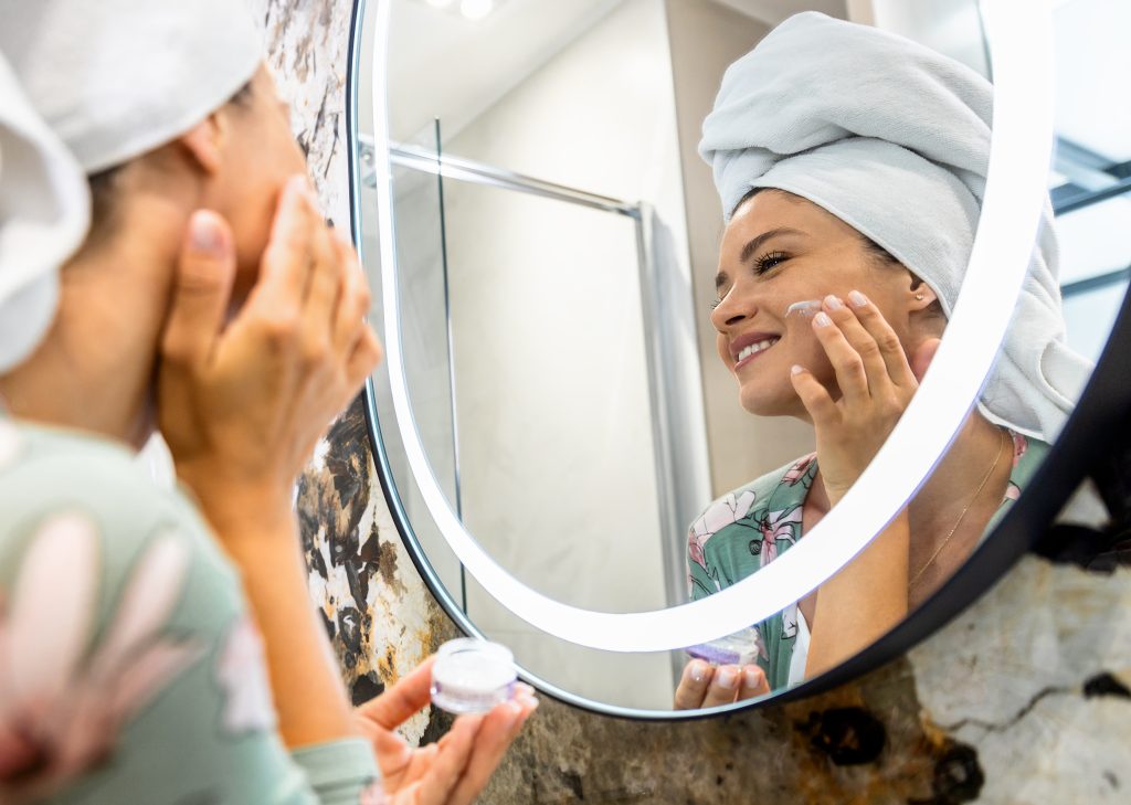 Smiling woman applies facial cream in her bathroom as part of her healthy morning routine.