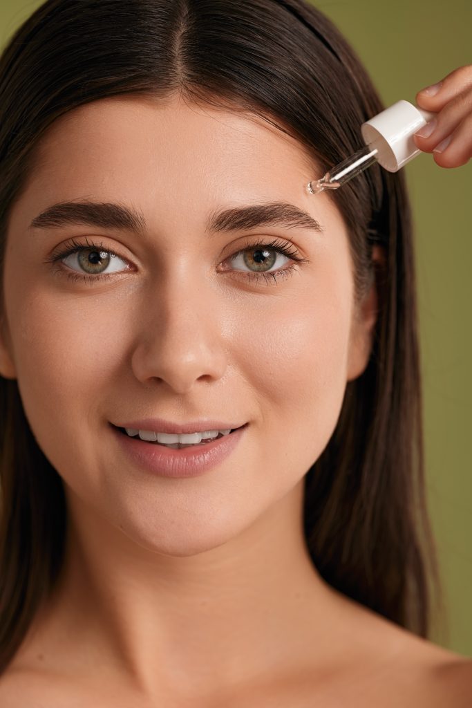 Positive young female millennial with long dark hair and perfect skin smiling and looking at camera, while applying cosmetic oil serum with dropper on face during skin care treatment