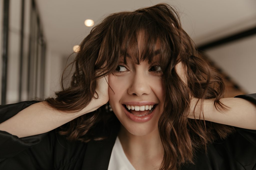 Surprised happy brown-eyed brunette woman in great mood ruffles hair. Charming pretty girl in black jacket smiles. Low-Maintenance Timeless Hairstyles.
