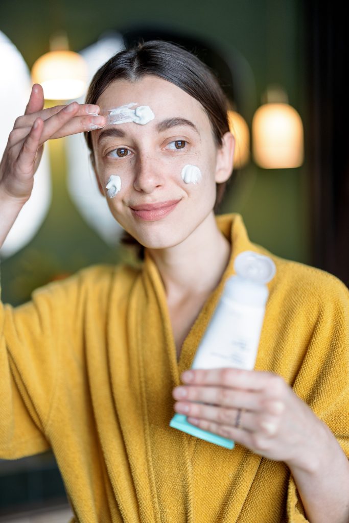 Slugging Skincare. Woman in yellow bathrobe applies moisturizer on her face. holding tube of cream in hand, having some morning beauty procedures at home