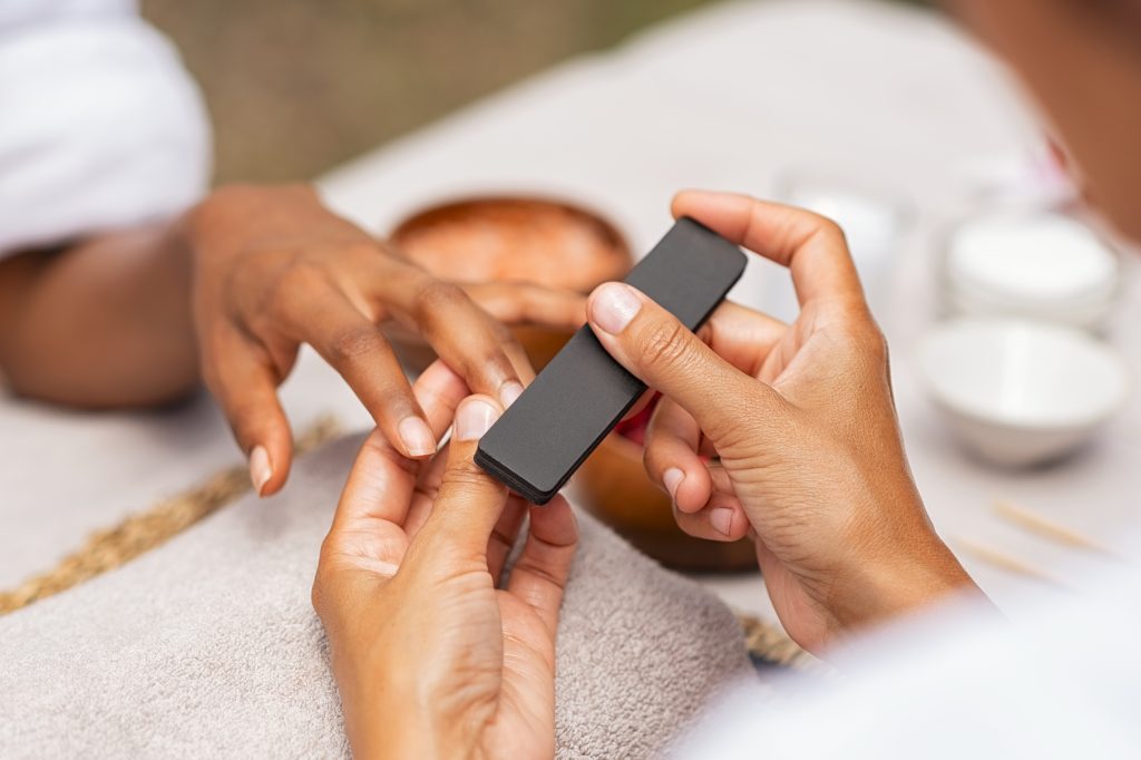 Nail Recovery After Gel - Restore Strength & Hydration Naturally. Close up of african hands of a qualified manicurist filing the nails of a young woman. Hands during manicure care session. Detail of a girl in a nail salon receiving manicure.