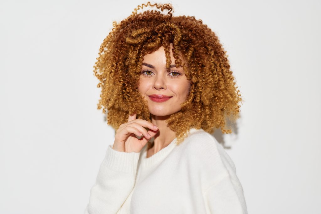A smiling woman with voluminous, curly hair is wearing a cozy white sweater The image features a simple white background, emphasizing her cheerful demeanor and natural beauty Perfect for concepts a