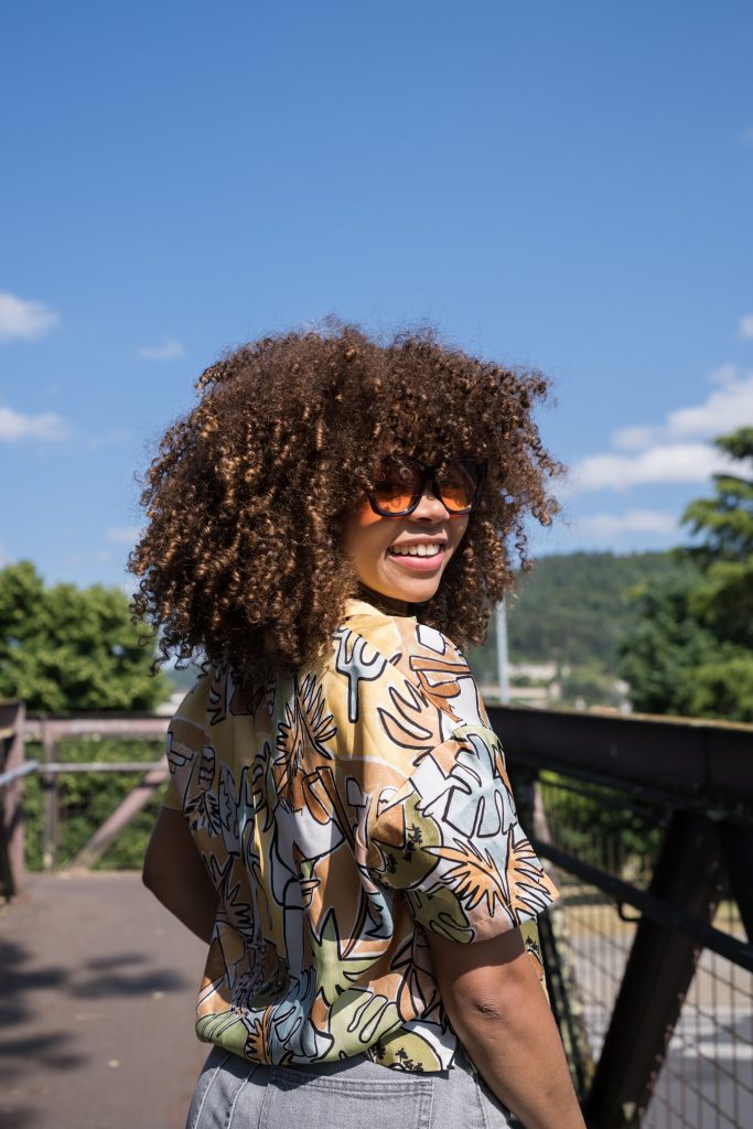 Young woman with curly hair and vibrant orange sunglasses smiling while glancing over her shoulder, walking confidently on a bridge under the bright sun on a cheerful summer day