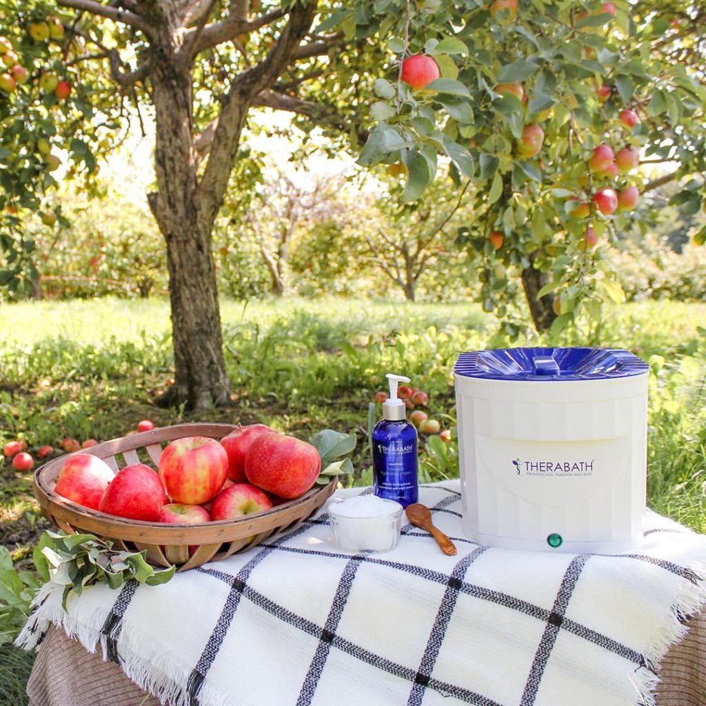A white Therabath paraffin wax bath with a blue lid sits on a checkered tablecloth in a sunlit apple orchard. Surrounding it are spa items including a blue pump bottle, a small white container, and a wooden spoon. A basket of red apples adds a natural touch, blending wellness and nature in a serene outdoor setting.