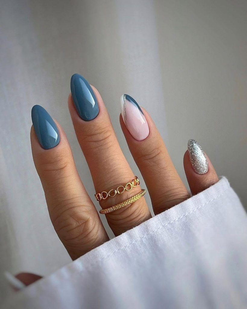 Close-up of a hand modeling a set of medium-long Almond nail shapes with a glossy, polished finish. The design features a bold dusty blue color, an accent nail with a chic minimalist French tip outlined in silver, and a second accent nail covered in sparkly silver glitter. This image visually represents a trendy, high-impact nail shape for teenagers looking for elegant yet easy DIY nail art at home.