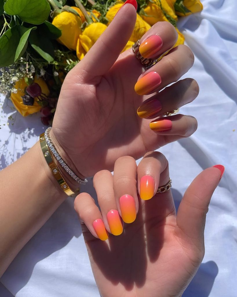 Two hands featuring nails filed into the Round and Oval nail shapes, which are easy for teenagers to try at home. The nails have a vibrant summer manicure with a pink and yellow ombré gradient, transitioning from hot pink near the cuticle to bright yellow at the tip. This photo emphasizes how classic nail shapes are the perfect canvas for bold and trendy DIY nail colors.