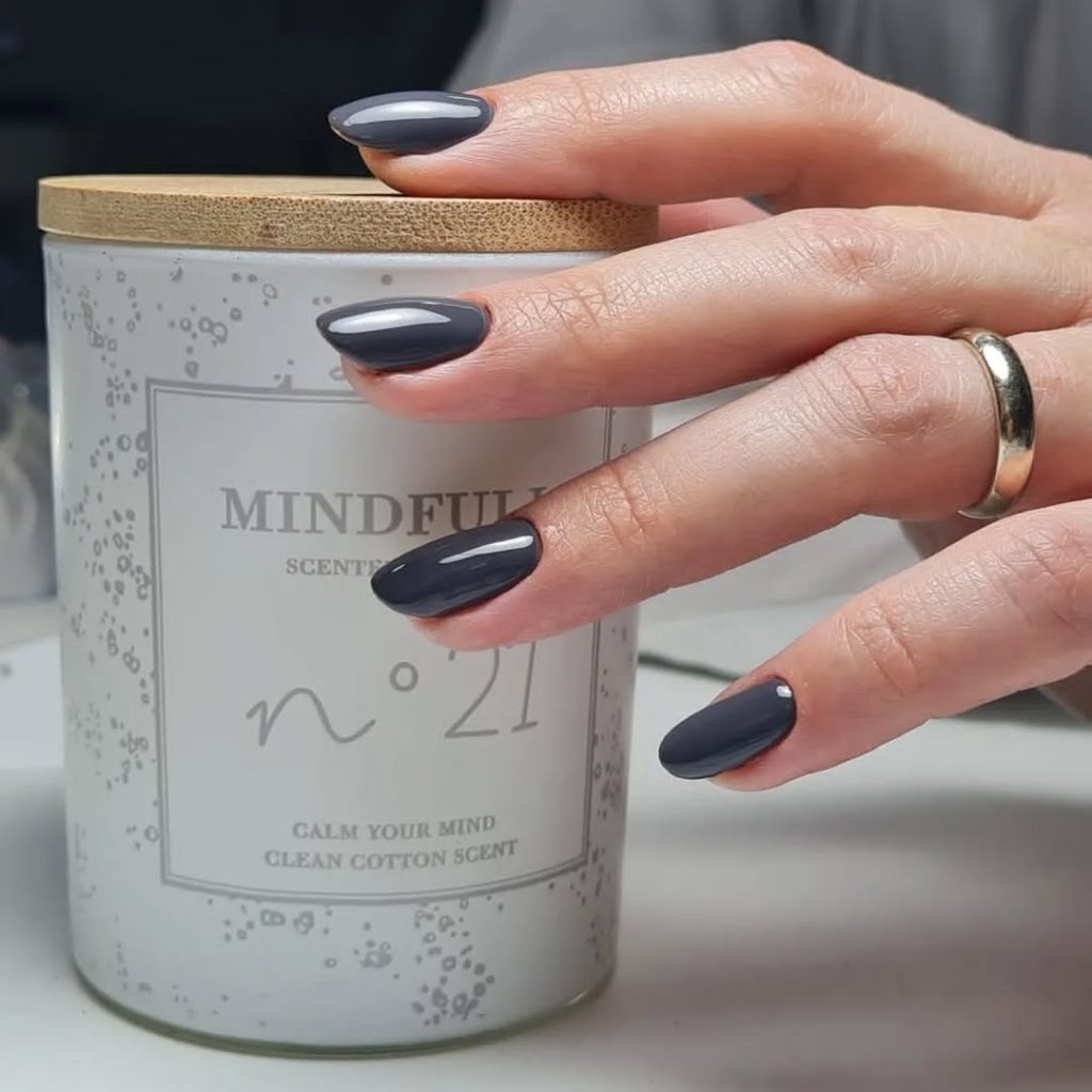 Close-up of a hand with short Oval nail shapes, filed for maximum durability and a neat, clean look. The nails are painted in a high-shine, glossy dark grey or charcoal polish. This image showcases a popular, low-maintenance nail shape for teenagers who prefer a neat, polished look but need a practical style that won't break while doing at-home manicures. The short length makes it an easy-to-maintain nail shape.