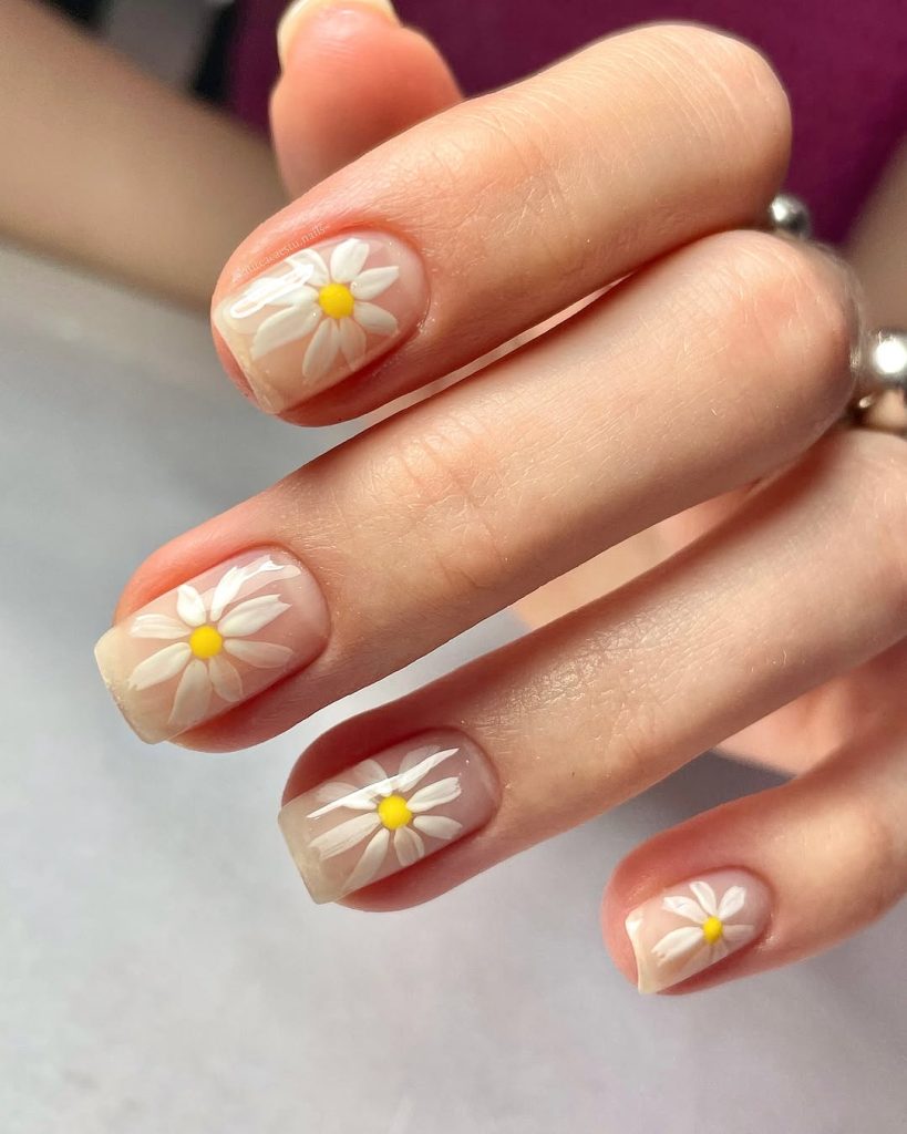 Close-up of a hand featuring short Squoval nail shapes, ideal for high durability and minimal breakage. The nails have a clear, milky base and feature popular white daisy flower nail art with yellow centers, a cute and easy design for teenagers to try at home. The squoval shape is one of the easiest nail shapes to maintain while still allowing for fun, detailed nail art.