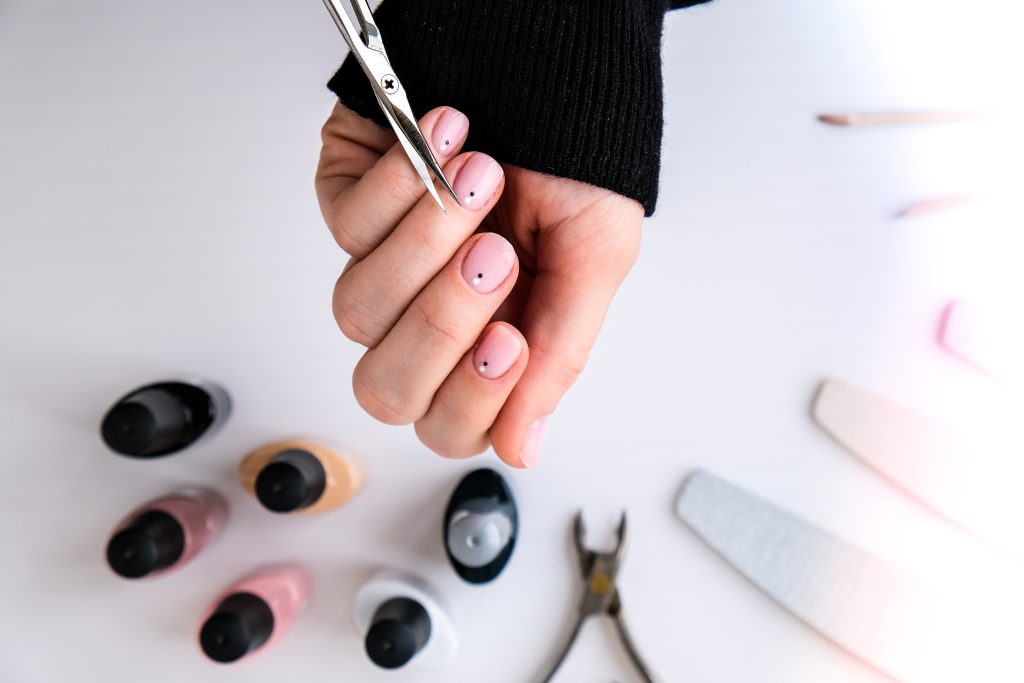 Overhead view of a hand with short Round nail shapes and a simple pale pink manicure with a black dot, demonstrating a teenager performing a DIY nail trim at home using small scissors. The surrounding table features essential manicure tools, including several bottles of nail polish, a buffer, and cuticle nippers. This image highlights the practicality of the Round nail shape and the importance of using the right tools for shaping natural nails.