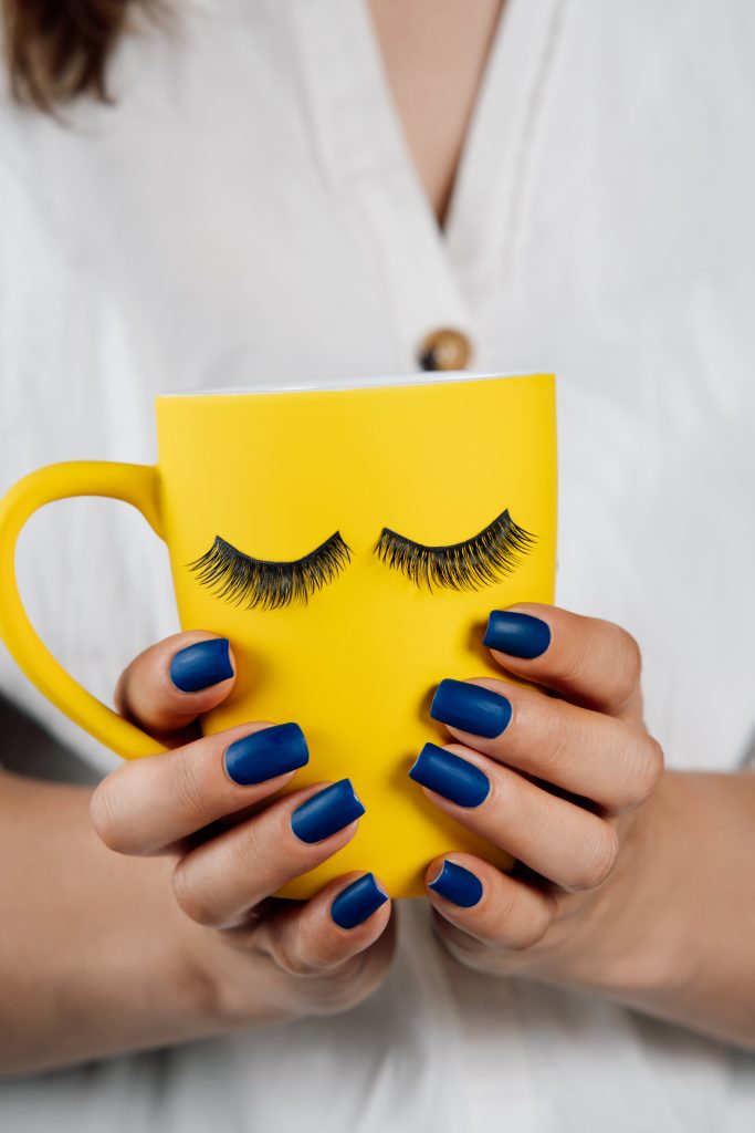 2025 Nail Polish Shades. Yellow cup of beverage with black fake eyelashes sticked on outside in hands of unrecognizable lady with blue manicure. Lashmaker holding mug with material for lash extension procedure. Vertical.