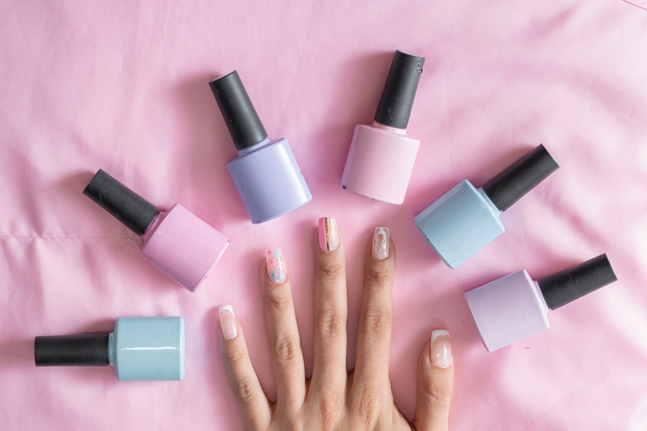 Detailed view of a woman's hand with painted nails surrounded by colored nail polish on a pink background. Gel nails last. Gel nail polish maintenance.