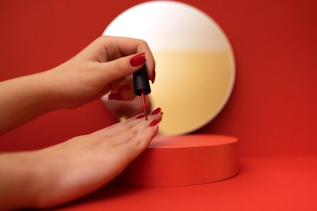 Young caucasian girl put her hands on a red podium and paints her nails with red polish. A round mirror with gold glitter on the background. Close up. Copy space.