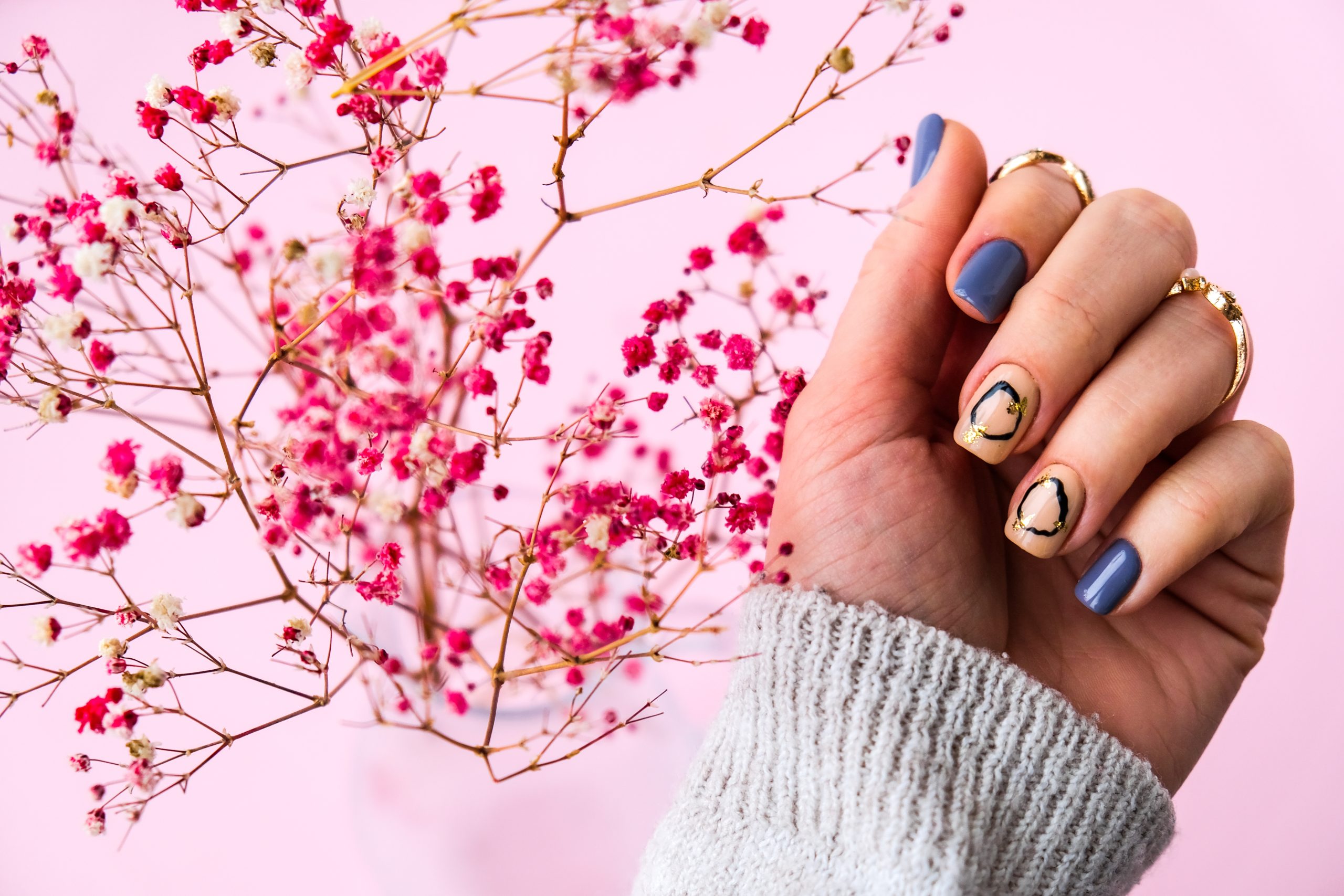 A hand showcasing a trendy 2026 minimalist manicure with short square nails. The design features a mix of dusty blue glossy polish and nude-toned accent nails decorated with delicate black circular fine-line art and tiny gold flake details, held against a backdrop of pink dried flowers.