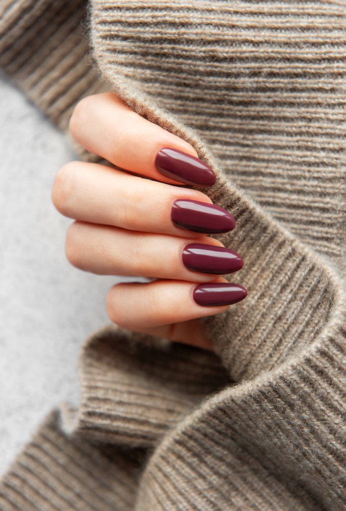 2026 color trends. A hand featuring elegant burgundy almond-shaped nails rests against a soft, knitted sweater, showcasing a trendy fall look that combines fashion and comfort.