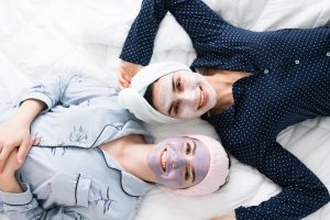 Portrait of two attractive women friends lying in bed and waiting for her face masks to clean their skin. Best friends feeling happy and relaxed in a slumber party after doing their daily skincare routine