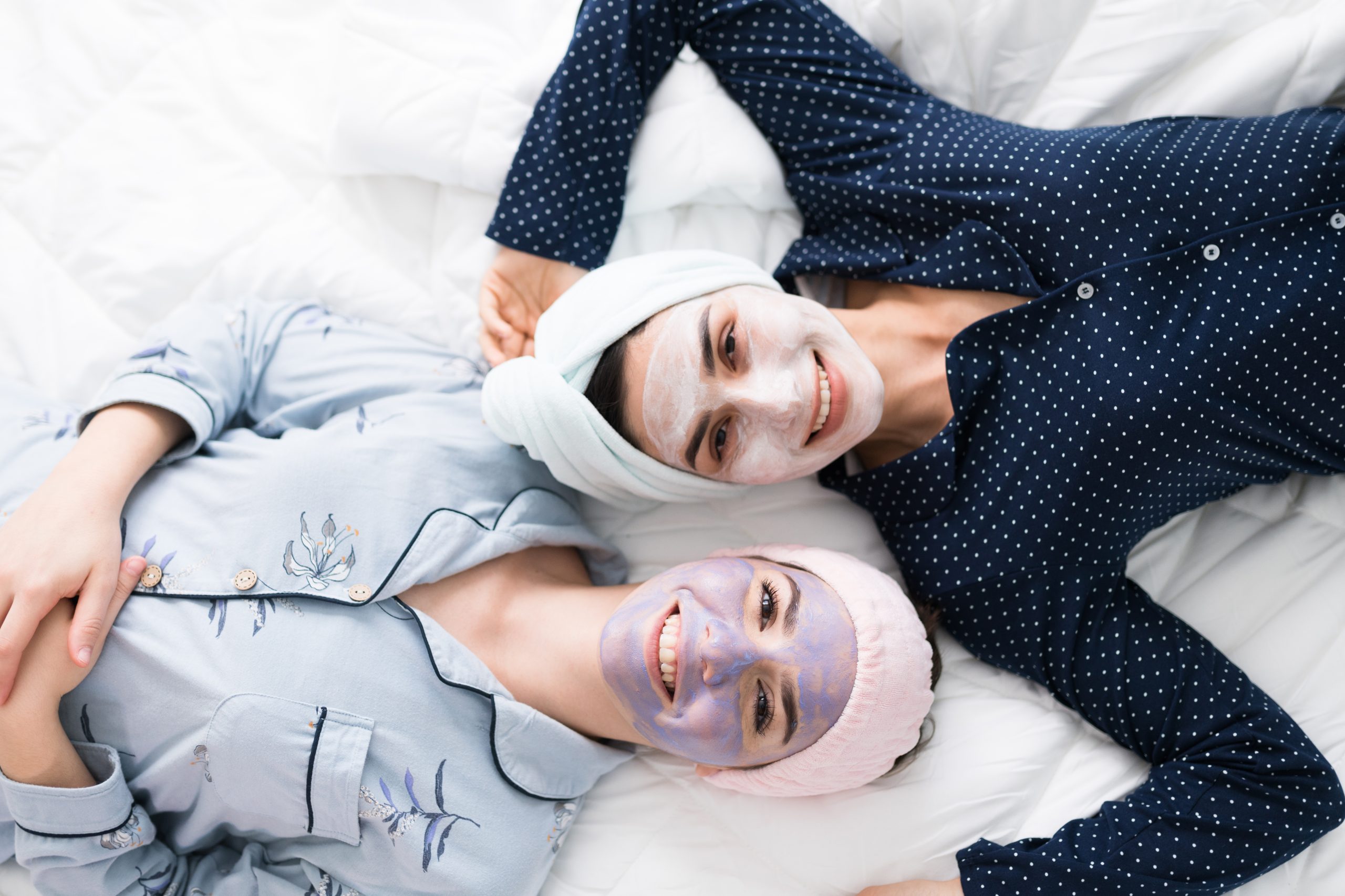 Portrait of two attractive women friends lying in bed and waiting for her face masks to clean their skin. Best friends feeling happy and relaxed in a slumber party after doing their daily skincare routine