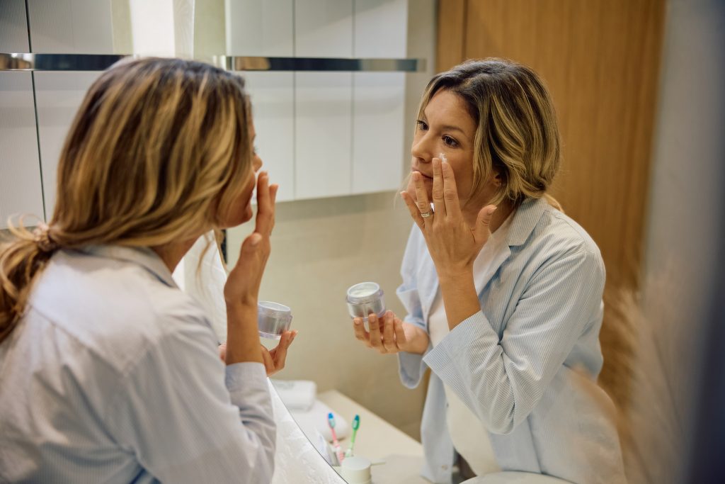 Mid adult woman using face moisturizer in front of a mirror.