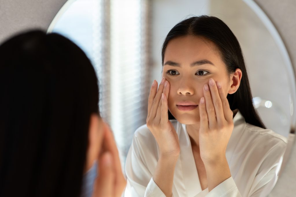 Mirror reflection of upset young asian woman in white bathrobe looking at mirror and touching her cheeks, having oily or dry skin problem, night skincare routine.