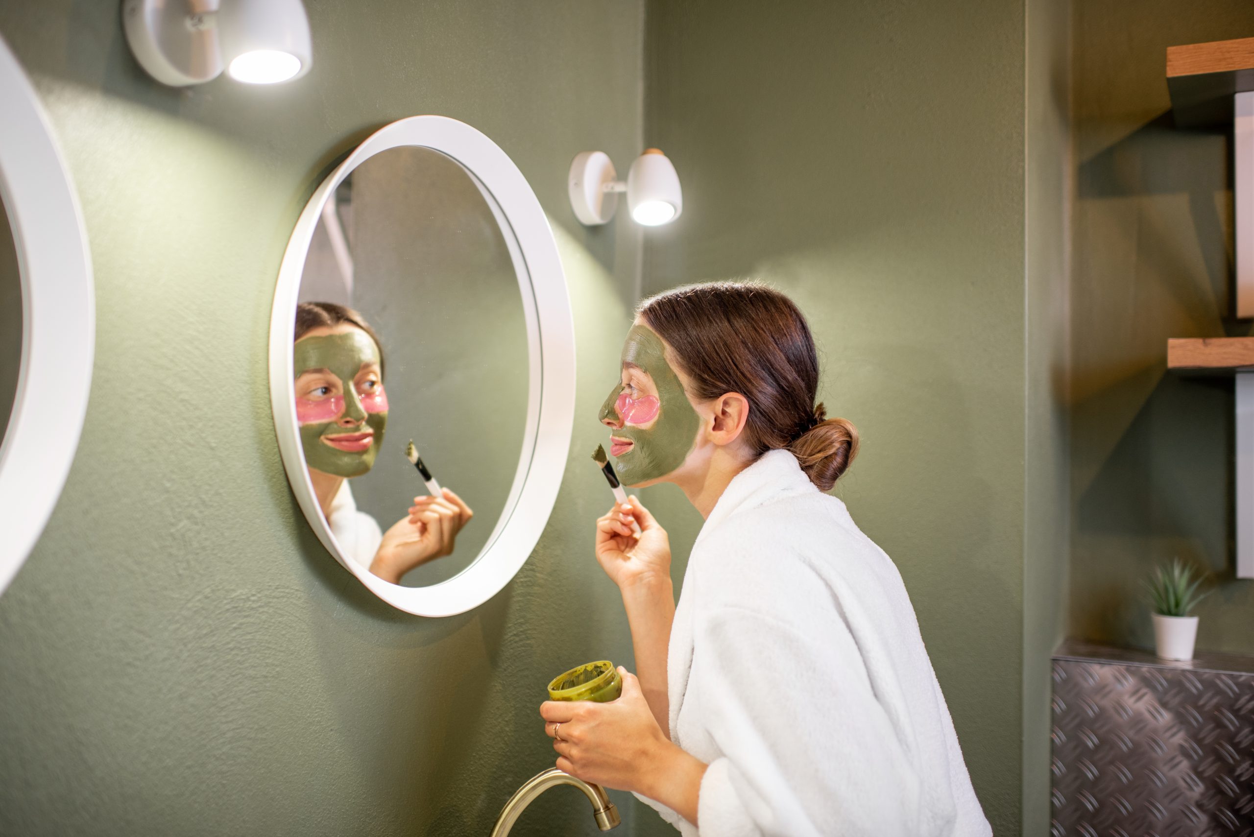 Night skincare routine, Young woman in bathrobe applying green mask on her face looking into the mirror in the modern green bathroom