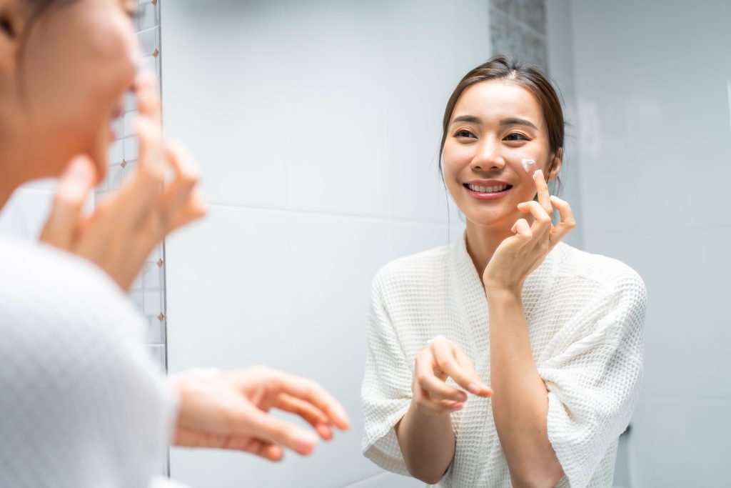 Asian beautiful woman looking at the mirror after apply lotion on face. Attractive young girl in bathrobe sitting on bed in bedroom, feel happy and proud for her healthy beauty treatment and daily skincare routine