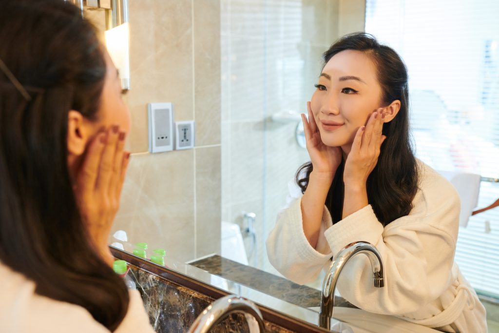 Satisfied young Asian woman in bathrobe standing in front of mirror in bathroom and washing face