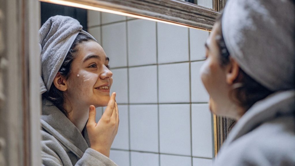 Teenager with towel-wrapped hair gently applies face cream in front of a mirror. Skincare-focused moment emphasizing dermatologist-recommended routines, natural glow, and youthful skincare habits.